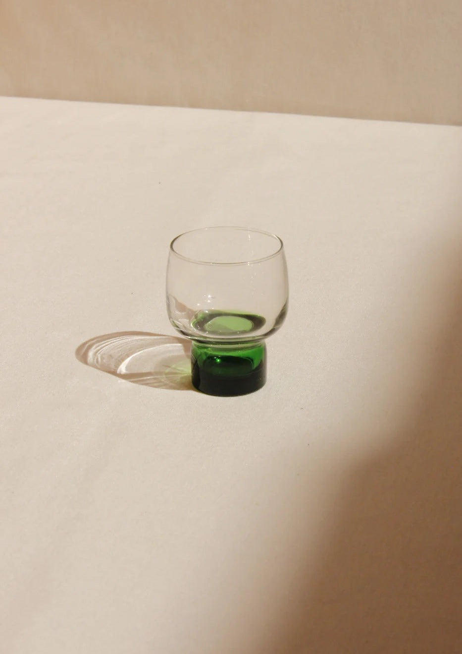 A small Set of French Mid-Century Glasses from Boga Avante Shop with a round base and a long, elegant shadow cast on a plain, slightly textured surface. The glass contains a small amount of green liquid.