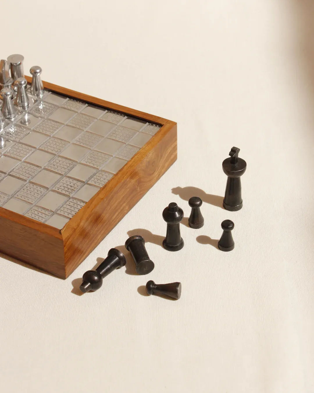 The Boga Avante Shop’s Wood and Metal Chessboard boasts a collectible wooden design with a silver metallic grid, elegantly displayed on a light surface. Black chess pieces such as pawns and the queen are artfully scattered, with side lighting enhancing its luxurious interior appeal.