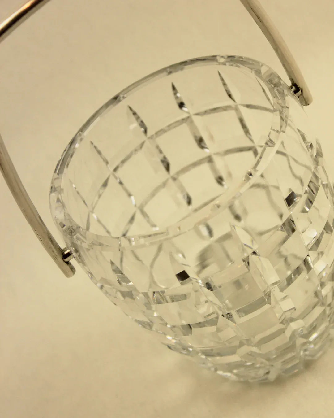 A close-up image showcases the Crystal Cubitera from Boga Avante Shop, a transparent, intricately patterned glass ice bucket with a brass handle. The luxury accessory is empty, and the photograph captures it from a slightly elevated angle, emphasizing the geometric design of the glass.