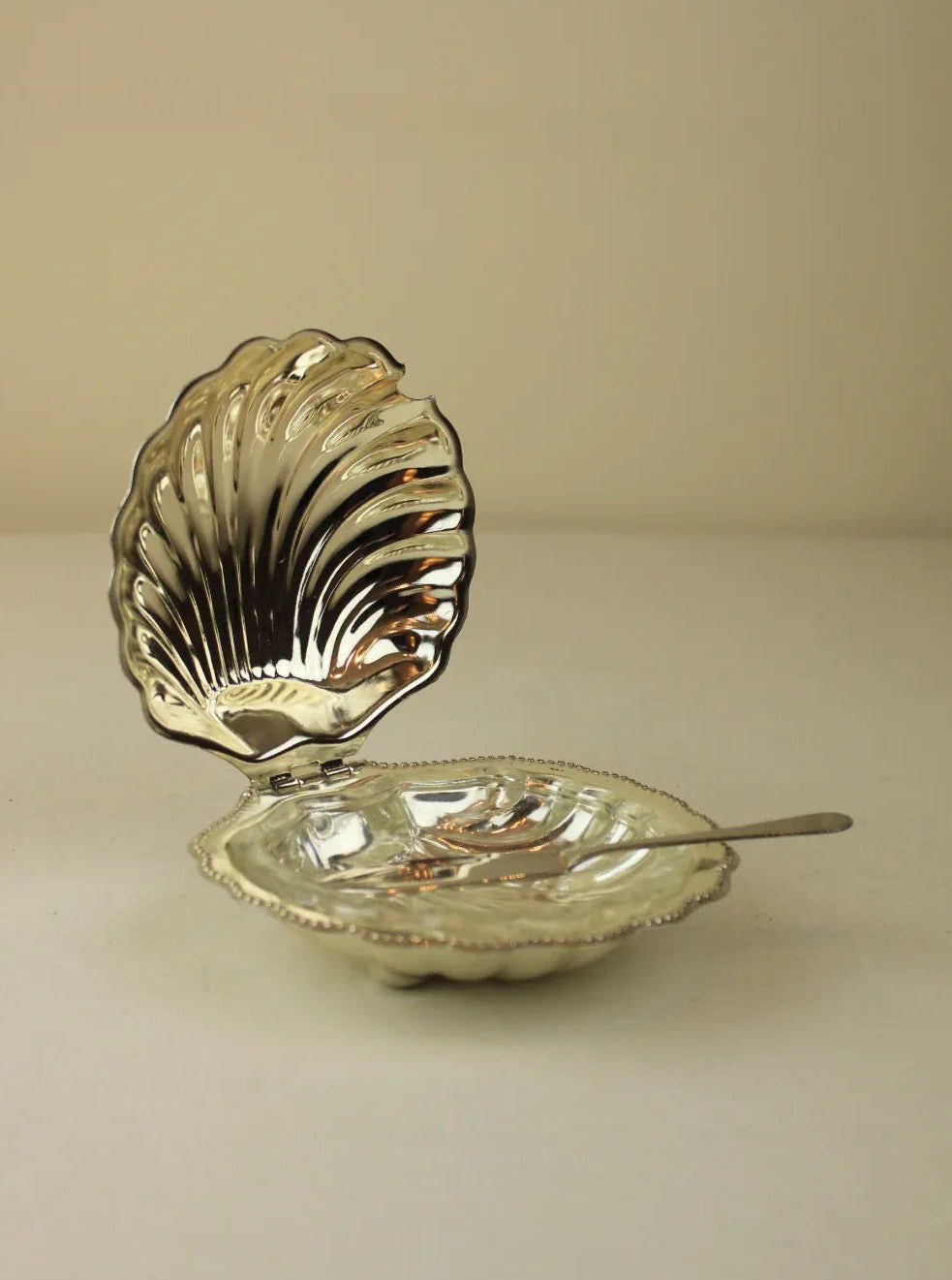 A polished, silver Shell Shaped Butter Dish from Boga Avante Shop features a hinged lid that opens to reveal an empty interior. Accompanied by a decorative butter knife, the reflective surfaces of both items highlight their intricate design and craftsmanship.