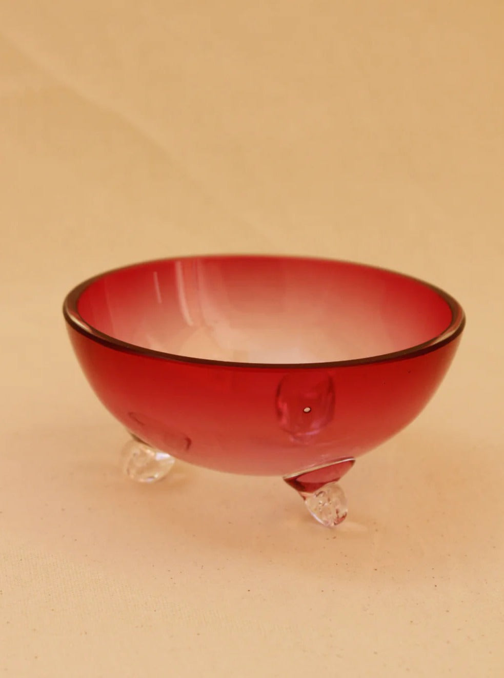 A pink Murano bowl from Boga Avante Shop with a smooth, glossy finish sits on three small transparent feet. Handcrafted with meticulous detail, it exudes a vintage allure. The bowl is slightly opaque, featuring a gradient effect from darker pink at the top edge to a lighter shade toward the bottom. The background is a soft beige color.