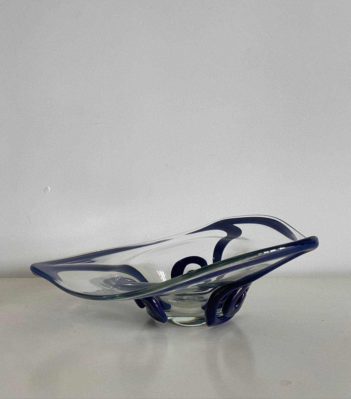 A modern, decorative Les Objets Amsterdam glass bowl with blue swirl designs, showcasing exquisite craftsmanship, resting on a plain white surface and backed by a white wall.