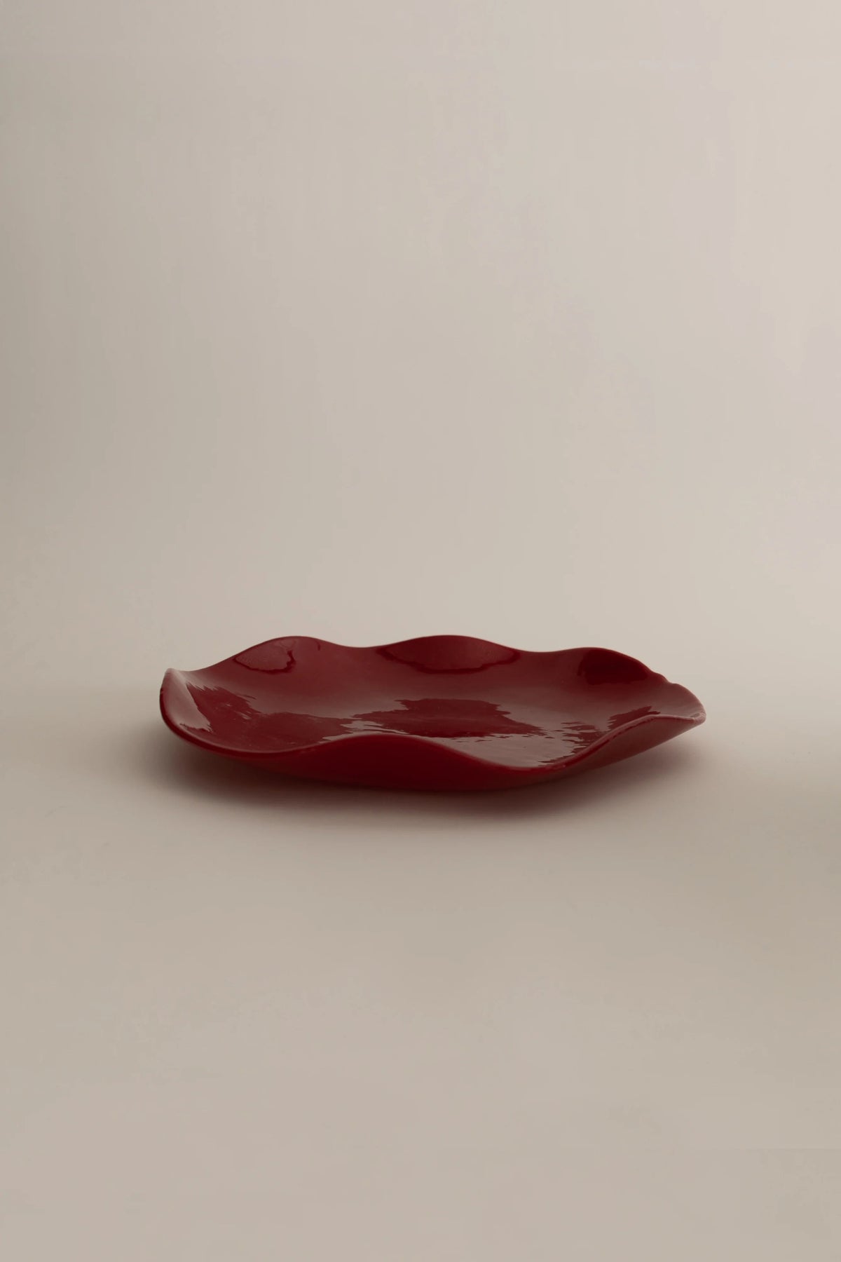 A glossy, irregularly shaped artisanal Sophie Lou Jacobsen Petal Plate - Large on a neutral beige background.