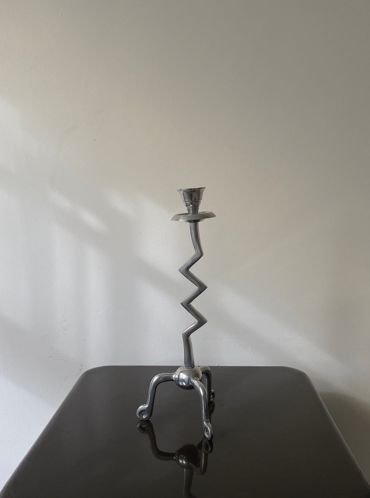 The Les Objets Amsterdam Vintage Zigzag Candle Holder, with its sculptural metal design and curved base, stands on a dark, glossy surface against a plain wall, enhancing the luxury interior appeal.