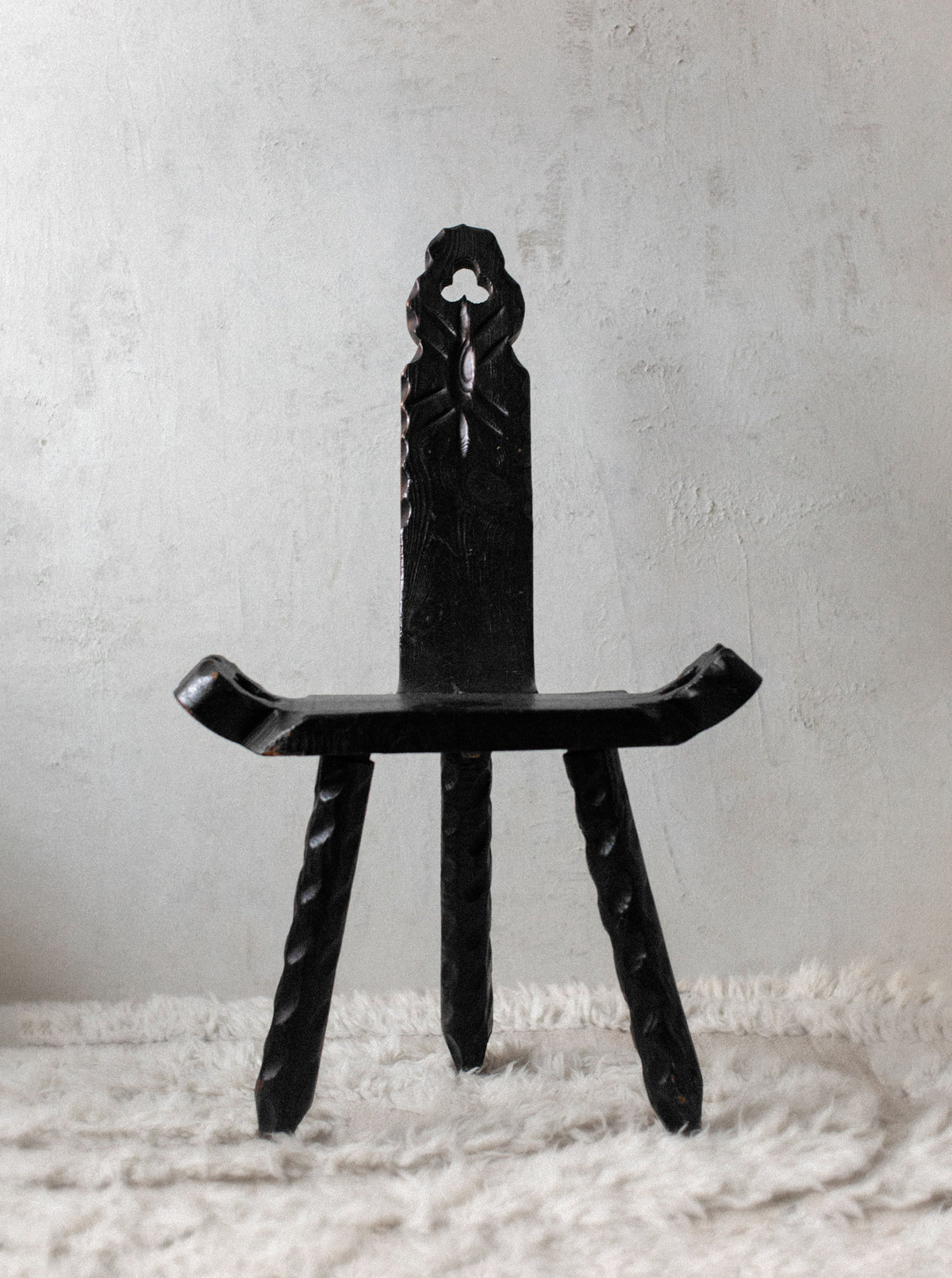 A black, ornate Out For Lunch Brutalist chair with intricate carvings, including a figure at the top of the backrest, stands on a rugged textured rug against a plain white wall.