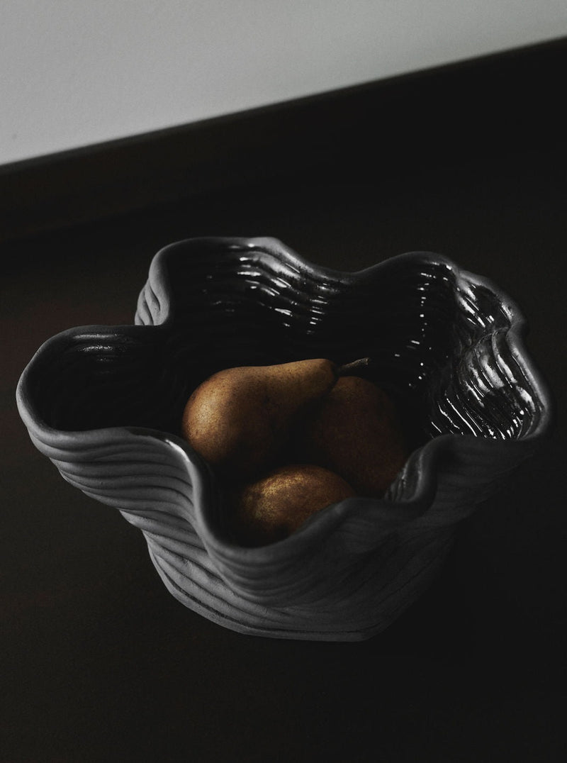 The 2222Studio Wobbly Bowl Black, with its wavy rim, cradles three pears in a contemporary design. In moody lighting, shadows enhance the texture of both the ceramic bowl and the fruit's surface.