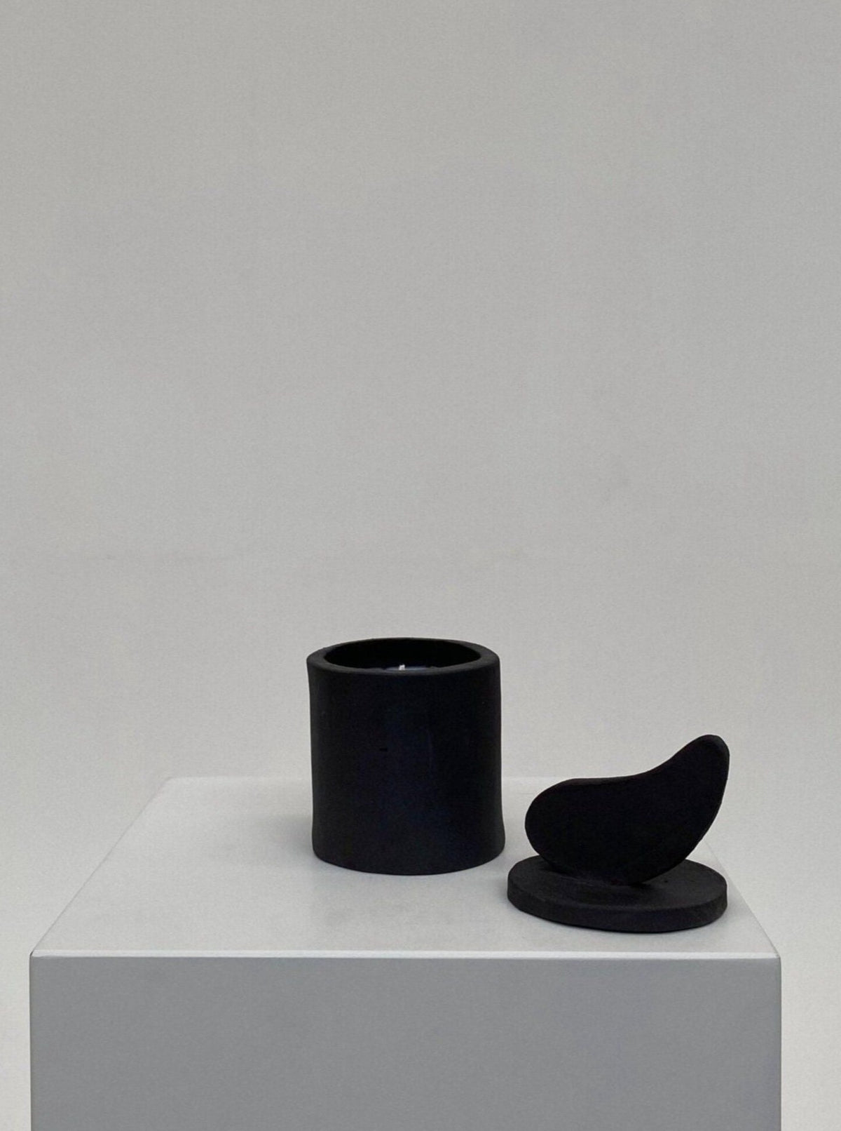 A minimalist display featuring a black cylindrical Sculptural Scented Candle No. 35 holder and a black sculptural object resembling a bird, both placed on a white pedestal against a light gray background.