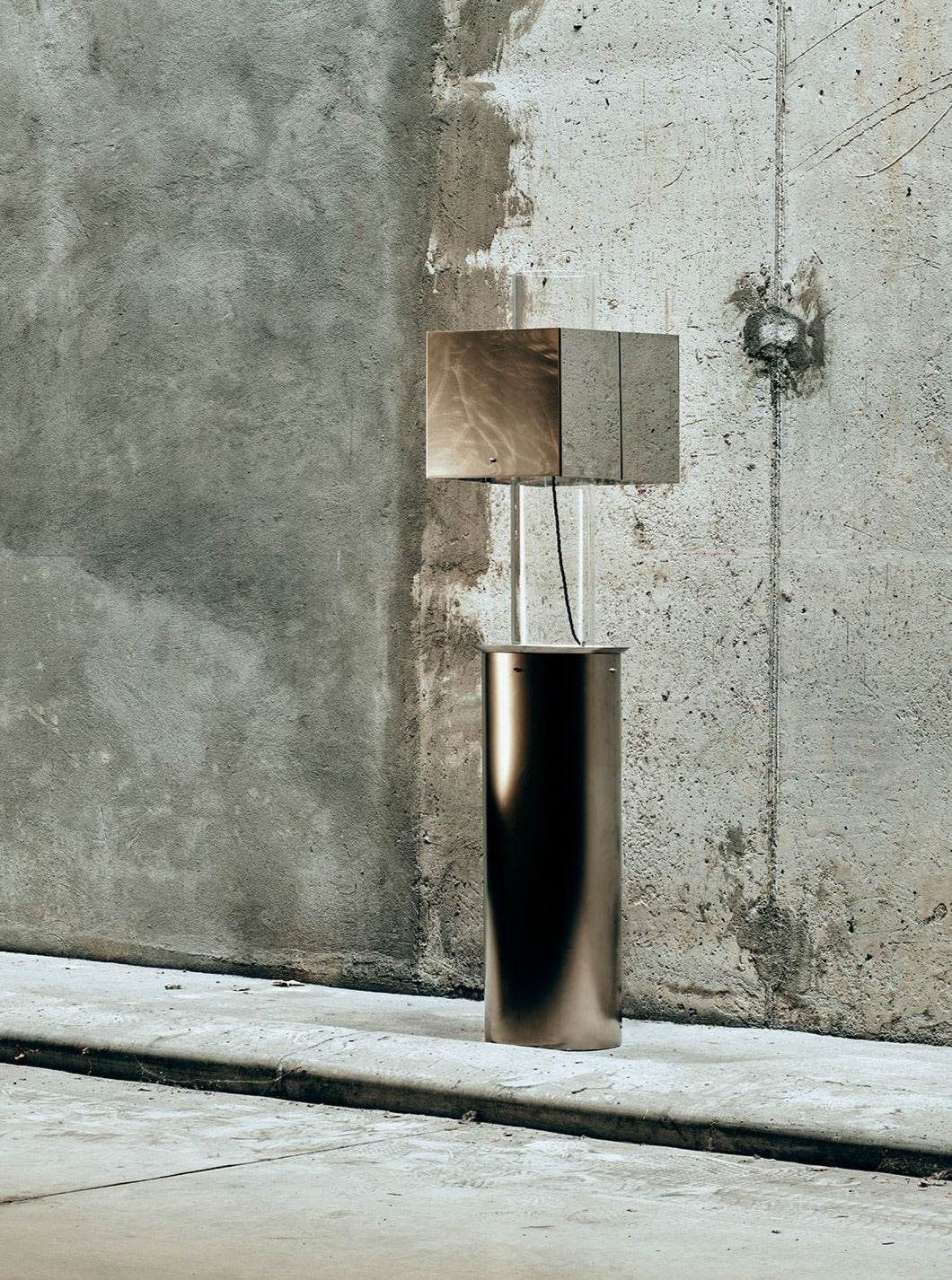 The Metal Floating Table Lamp by Brajak Vitberg, with its metallic cube and electrical wires on a cylindrical pedestal against a textured concrete wall, exudes a minimalist aesthetic and industrial design, adding sophistication to any luxury interior decor.