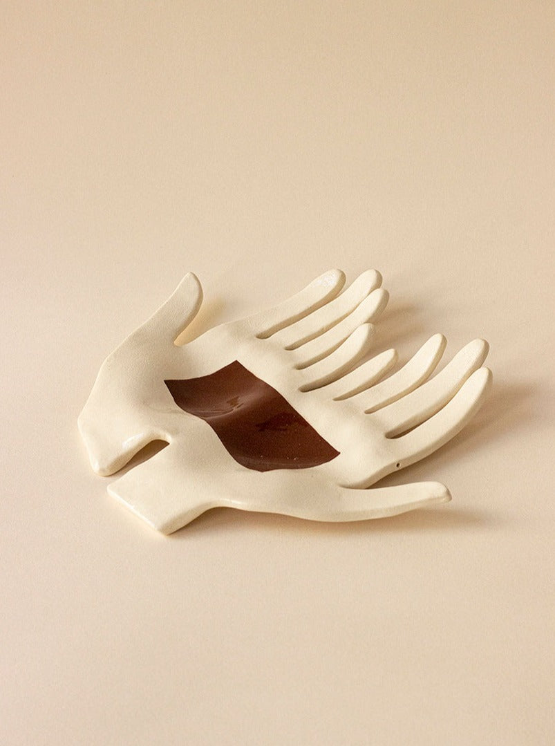 The Main Edition's Main Double "What binds us" Brown Bond is a handcrafted ceramic sculpture featuring two open hands with a central rectangular cutout. Its contemporary design and elegance make it an exquisite addition to any space, accentuating its artistic charm against a neutral backdrop.